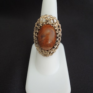 May include: A vintage ring with a large, oval, brown stone set in a gold-toned filigree frame. The ring is displayed on a white ring holder against a black background, showcasing its intricate design and warm color palette.