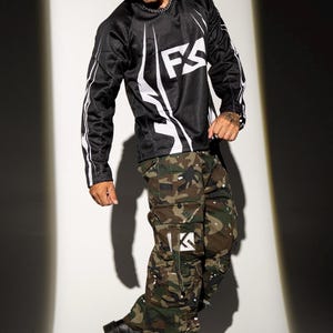 May include: Black long-sleeved mesh shirt with white accents and the letters "FS" in white. The person is wearing camouflage cargo pants and black boots. The pants have white paint splatters and the letter "K" in white.