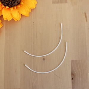 May include: Two white, curved bra underwires are positioned on a light wood surface. Two yellow sunflowers are in the upper left corner. The underwires are made of a flexible material.