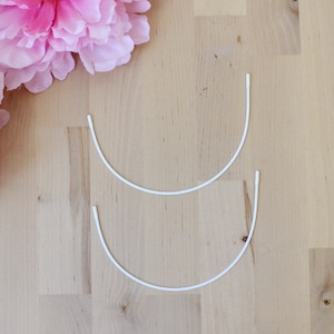 May include: Two white bra underwires are arranged on a light wood surface, with two large pink peony flowers in the upper left corner. The underwires are curved and have a rounded shape.