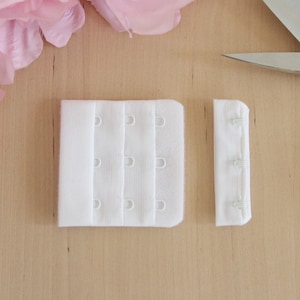 May include: Two white bra extenders with hook and eye closures, one with three rows of hooks and the other with three eyes. A pair of scissors and pink flower petals are in the background.