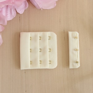 May include: Two ivory bra hook and eye closures with gold-colored metal hardware. One closure has three rows of hooks and eyes, and the other has three eyes. A pair of scissors is on the right side of the image.