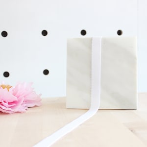 May include: A white marble block with a white ribbon wrapped around it. Pink peonies are on the left side of the block. The background is a white wall with black dots. The objects are on a light wood surface.