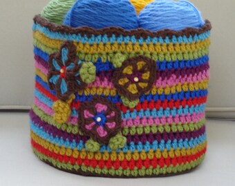 May include: A colorful crocheted yarn basket with a striped pattern and flower embellishments. The basket is filled with balls of yarn in various colors.