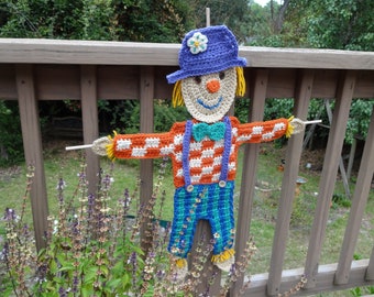 May include: A crocheted scarecrow with a purple and white hat, orange and white checkered shirt, and blue and green pants. The scarecrow is standing on a wooden deck with green plants in the background.