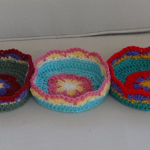 May include: Three crocheted bowls with colorful flower designs. The bowls are green, blue, and red with yellow, white, and purple accents.