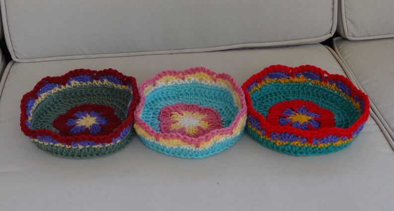 May include: Three crocheted bowls with colorful flower designs. The bowls are green, blue, and red with yellow, white, and purple accents.