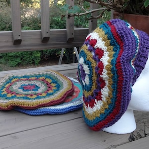 Op de afbeelding: Een kleurrijke gehaakte beanie met een paars, rood, wit en gouden patroon. De beanie zit op een wit mannequin hoofd.