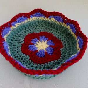 May include: A crocheted bowl with a scalloped edge. The bowl is made with green, red, blue, and yellow yarn. The center of the bowl has a flower design.