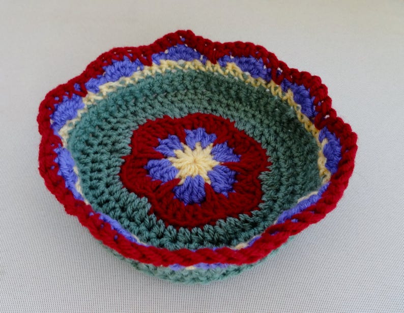 May include: A crocheted bowl with a scalloped edge. The bowl is made with green, red, blue, and yellow yarn. The center of the bowl has a flower design.