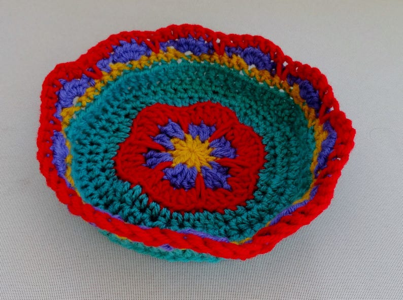May include: A crocheted bowl with a colorful flower design. The bowl is made with red, yellow, blue, and green yarn.
