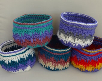 May include: Five crocheted baskets in different colors and patterns. The baskets are made with a variety of yarn colors, including purple, teal, red, white, and yellow.