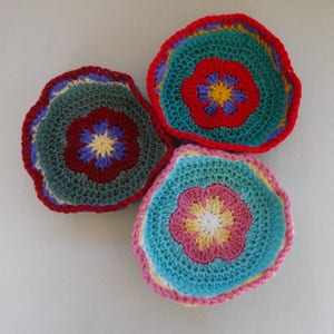May include: Three crocheted bowls with colorful flower designs. The bowls are teal, green, and pink with yellow, red, and white accents.