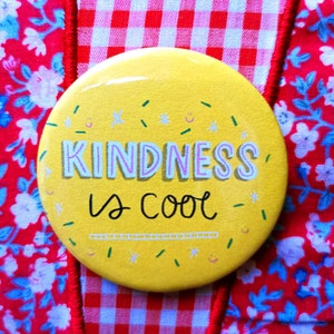 Könnte beinhalten: Ein gelber Knopf mit dem Text "KINDNESS is COOL" in rosa und weißen Buchstaben. Der Knopf ist von einem rot-weiß karierten Muster umgeben.