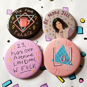 May include: Four round pink, black, and gray button pins with different designs. The first pin has a pink coffee mug with the text "The Pink Palace" in black. The second pin has a cartoon of a woman with the text "Be More Jill". The third pin has the text "23, Piss Off Avenue London, W FUCK". The fourth pin has a blue triangle with the text "La!" in white.