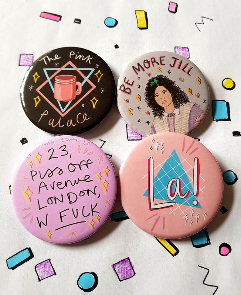 May include: Four round pink, black, and gray button pins with different designs. The first pin has a pink coffee mug with the text "The Pink Palace" in black. The second pin has a cartoon of a woman with the text "Be More Jill". The third pin has the text "23, Piss Off Avenue London, W FUCK". The fourth pin has a blue triangle with the text "La!" in white.
