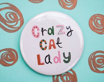 Könnte beinhalten: Ein runder Ansteckbutton mit weißem Hintergrund, der den Schriftzug "Crazy Cat Lady" in mehrfarbigen, stilisierten Buchstaben zeigt. Die Buchstaben sind in verschiedenen Rosa-, Grün-, Orange- und Schwarztönen mit Mustern gestaltet.