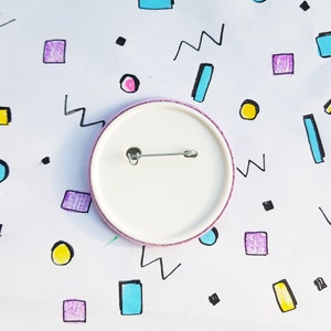 May include: The back of a white button with a pink border and a safety pin attached. The button is on a white background with colorful geometric shapes.