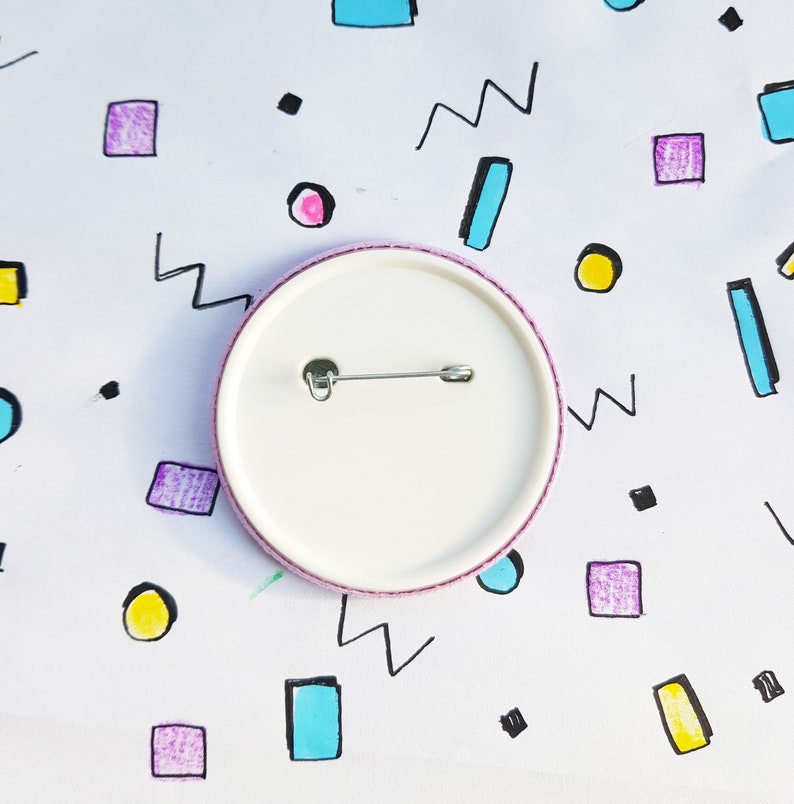 May include: The back of a white button with a pink border and a safety pin attached. The button is on a white background with colorful geometric shapes.