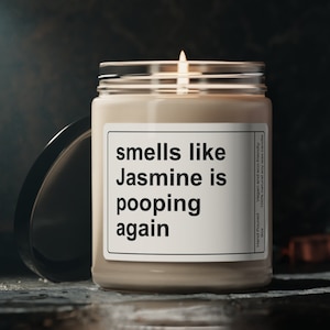 May include: A brown candle with a white label that reads "smells like Jasmine is pooping again". The candle is lit and the flame is visible.