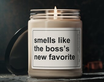 May include: A beige candle with a white label that reads "smells like the boss's new favourite". The candle is lit and the flame is visible.