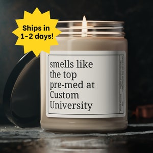Puede incluir: Una vela blanca en un tarro de vidrio transparente con una etiqueta que dice "smells like the top pre-med at Custom University". La vela está encendida y la llama es visible.