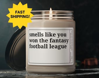 May include: A beige candle with a white label that reads "smells like you won the fantasy football league". The candle is in a glass jar with a black lid.