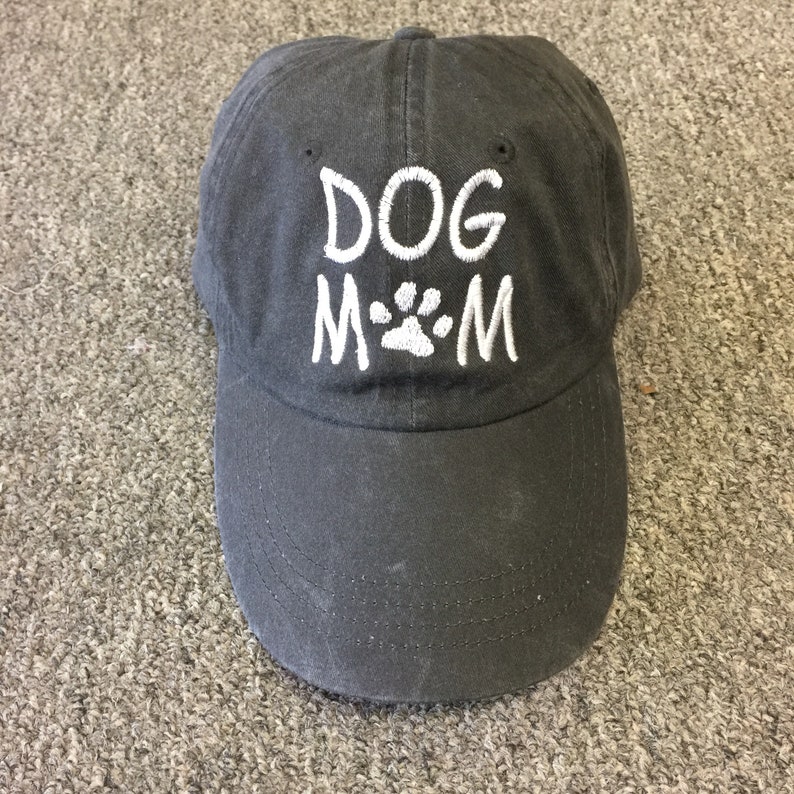 K&ouml;nnte beinhalten: Eine schwarze Baseballkappe mit einem wei&szlig;en gestickten Design, das "DOG M*M" mit einem Pfotenabdruck in der Mitte sagt.