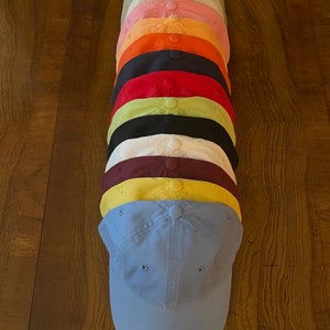 K&ouml;nnte beinhalten: Ein Stapel von 10 einfarbigen Baseballkappen in verschiedenen Farben, darunter hellgrau, rosa, orange, schwarz, rot, gr&uuml;n, wei&szlig;, weinrot, gelb und hellblau.