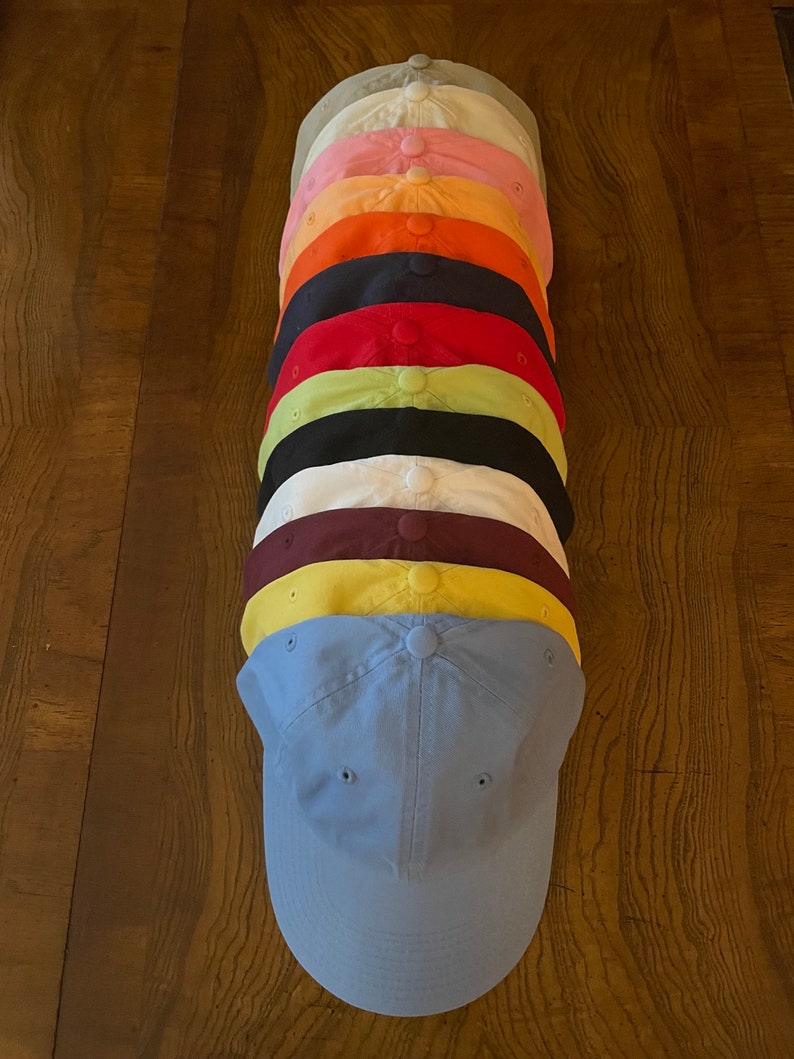 K&ouml;nnte beinhalten: Ein Stapel von 10 einfarbigen Baseballkappen in verschiedenen Farben, darunter hellgrau, rosa, orange, schwarz, rot, gr&uuml;n, wei&szlig;, weinrot, gelb und hellblau.