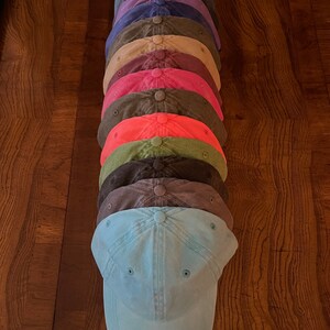 K&ouml;nnte beinhalten: Ein Stapel von zehn einfarbigen Baseballkappen in verschiedenen Farben, darunter Blau, Lila, Braun, Gr&uuml;n, Rot und Pink. Die Kappen sind in einem vertikalen Stapel angeordnet, wobei die unterste Kappe hellblau ist.