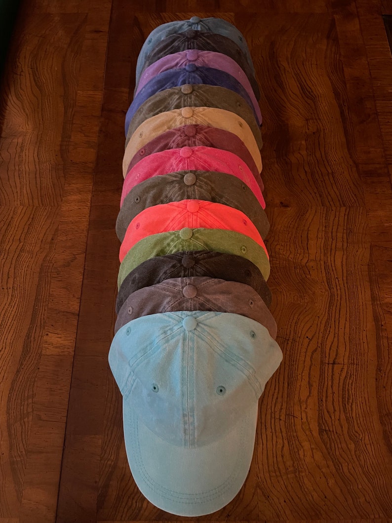 K&ouml;nnte beinhalten: Ein Stapel von zehn einfarbigen Baseballkappen in verschiedenen Farben, darunter Blau, Lila, Braun, Gr&uuml;n, Rot und Pink. Die Kappen sind in einem vertikalen Stapel angeordnet, wobei die unterste Kappe hellblau ist.