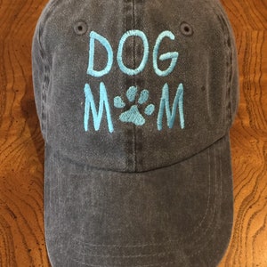 K&ouml;nnte beinhalten: Eine dunkelblaue Baseballkappe aus Denim mit den Worten "DOG MOM" in hellblau gestickt, in der Mitte ein Pfotenabdruck.