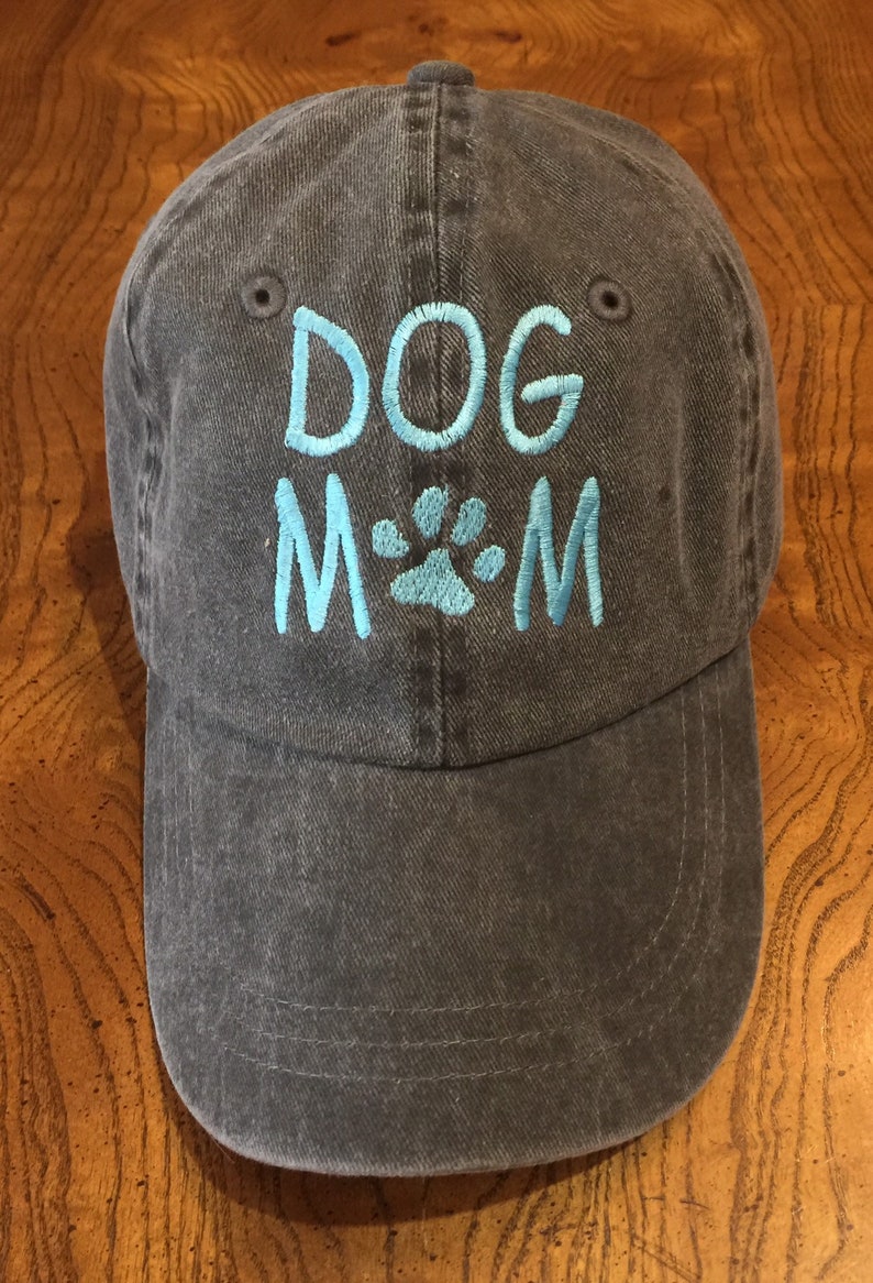 K&ouml;nnte beinhalten: Eine dunkelblaue Baseballkappe aus Denim mit den Worten "DOG MOM" in hellblau gestickt, in der Mitte ein Pfotenabdruck.