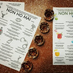 May include: A white card with black text lists a series of prompts for a drinking game. The prompts are written in Italian and include phrases like "Non ho mai..." and "Baciato due persone diverse nella stessa sera". The card is surrounded by small shot glasses filled with a dark liquid.