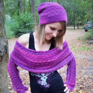 May include: A woman wearing a purple knitted hat and matching shawl over a black shirt with a floral design. She is wearing blue jeans.