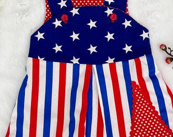 May include: A red, white, and blue sleeveless dress with a star print on the top and a striped print on the bottom. The dress has a red polka dot fabric on the back and a red bow headband with white pearls.