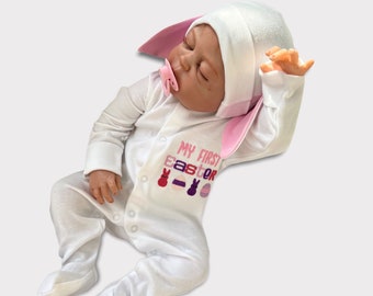 Könnte beinhalten: Ein weißer Baby-Strampler mit rosa Hasenohren und dem Text "My First Easter" in rosa, lila und roten Buchstaben. Der Strampler hat gestickte Ostern-Motive, darunter ein Hase, ein Ei und ein Cupcake.