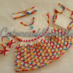 May include: A crocheted baby outfit set in white with colorful accents. The set includes a dress, a headband, booties, and a diaper cover. The dress has a colorful geometric pattern and is decorated with colorful buttons. The headband is decorated with colorful buttons. The booties are white with red bows. The diaper cover is white with colorful trim.