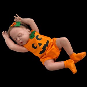 May include: A baby wearing an orange pumpkin costume with a green leaf and black jack-o'-lantern face. The baby is wearing a headband with a green leaf and orange flower.