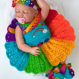 Puede incluir: Una mu&ntilde;eca beb&eacute; con un vestido de crochet arco&iacute;ris con una diadema y botines a juego. El vestido tiene un dise&ntilde;o de arco&iacute;ris con una nube blanca y un arco&iacute;ris en la parte delantera. La diadema est&aacute; hecha de flores de tela arco&iacute;ris.
