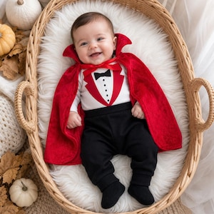 May include: A baby outfit consisting of a white long-sleeve shirt with a printed red tuxedo vest and black bow tie, paired with solid black pants and matching black booties. A vibrant red velvet cape with a high collar is worn over the outfit.