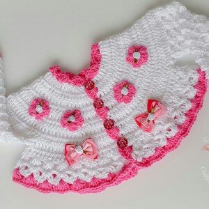 May include: White crocheted baby cardigan with pink trim and flower accents. The cardigan has a button closure and is decorated with pink bows.