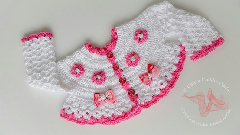 May include: White crocheted baby cardigan with pink trim and flower accents. The cardigan has a button closure and is decorated with pink bows.