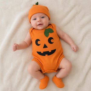 May include: An orange sleeveless bodysuit featuring a black jack-o'-lantern face print and a green fabric leaf accent on the chest, paired with a matching orange beanie hat with a small green stem and a pair of solid orange baby booties.