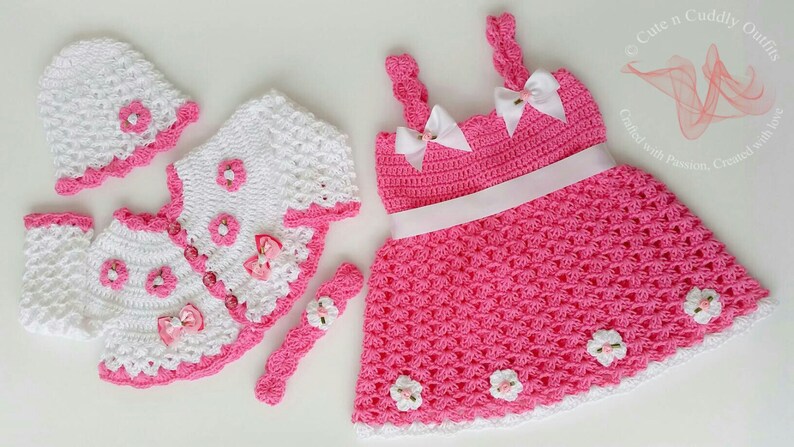 May include: A crocheted baby outfit set in pink and white. The set includes a dress, a cardigan, a hat, and a headband. The dress has a white ribbon around the waist and white crocheted flowers on the skirt. The cardigan has a white crocheted flower on the front. The hat has a white crocheted flower on the top. The headband has a white crocheted flower on the front.