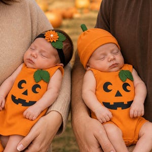 May include: Two infants wearing orange sleeveless bodysuits with black jack-o'-lantern face prints and green felt leaf accents. One infant wears a black headband with an orange fabric flower, while the other wears a matching orange beanie hat.
