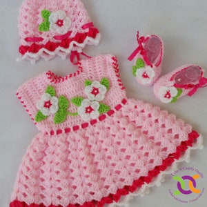 Op de afbeelding: Een roze gehaakte baby-outfit met witte en groene bloemenaccenten. De outfit bestaat uit een jurkje, een muts en slofjes. Het jurkje heeft een gegolfde zoom en een roze lint om de taille. De muts heeft een gegolfde rand en een witte bloem bovenop. De slofjes hebben een witte bloem aan de voorkant en een roze lint bovenop.