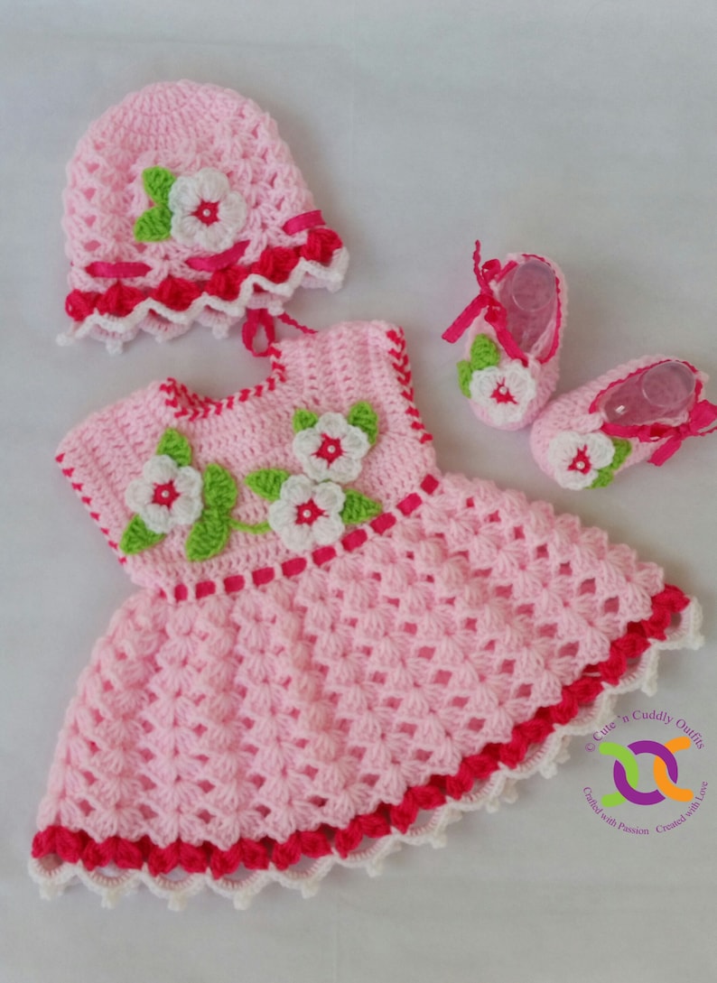 Op de afbeelding: Een roze gehaakte baby-outfit met witte en groene bloemenaccenten. De outfit bestaat uit een jurkje, een muts en slofjes. Het jurkje heeft een gegolfde zoom en een roze lint om de taille. De muts heeft een gegolfde rand en een witte bloem bovenop. De slofjes hebben een witte bloem aan de voorkant en een roze lint bovenop.