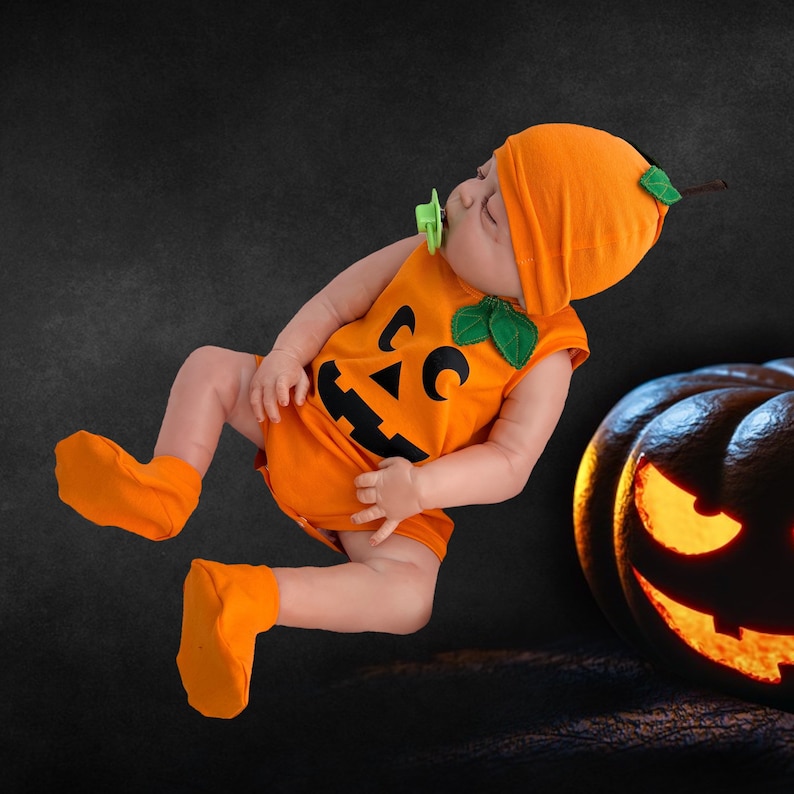 May include: A baby wearing an orange pumpkin costume with a jack-o'-lantern face. The costume has green leaves on the shoulders and a matching hat.